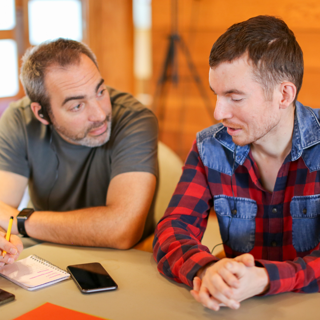 A white man with short greying hair and a young man with dark hair engaging in conversation and writing notes at the in-person Youth Leaders training.