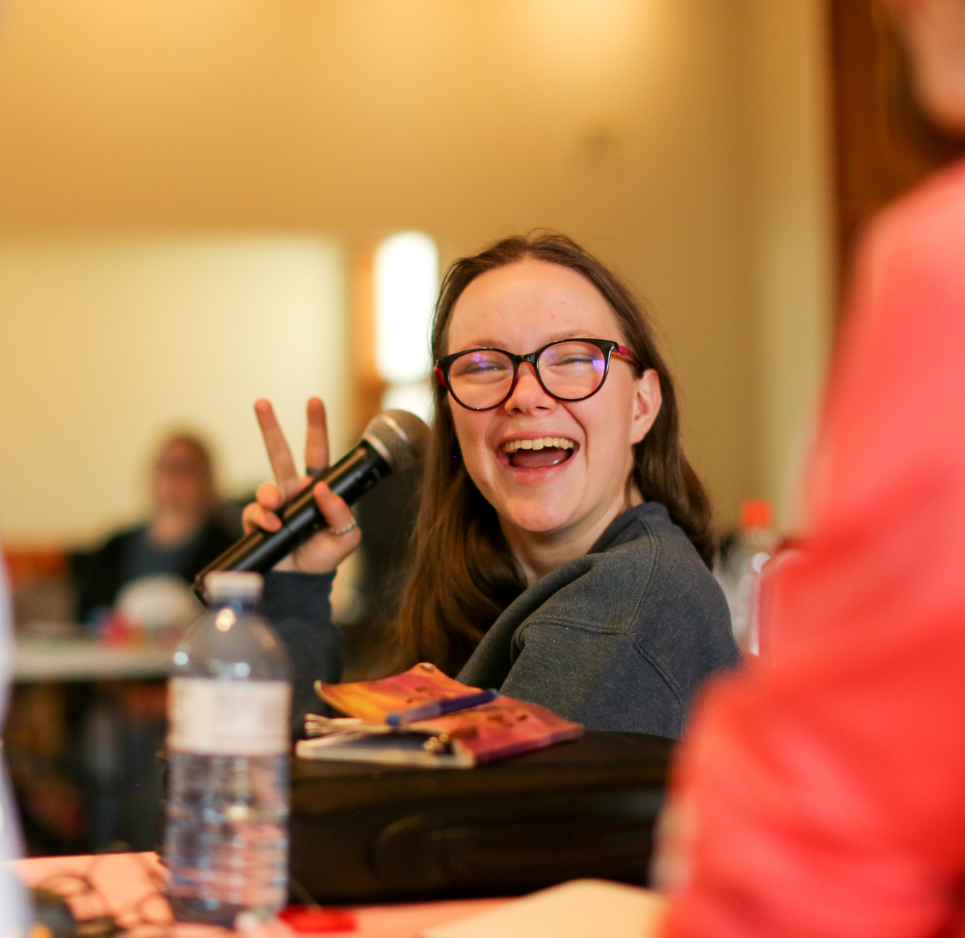 A young white woman with glasses laughing, holding up microphone and peace sign with her hands, at the Youth Leaders in person training.