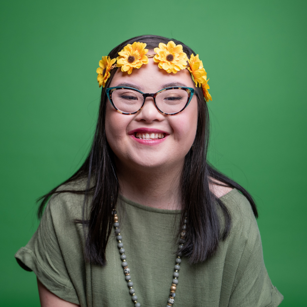 A smiling dark haired young woman with down syndrome wearing glasses and a yellow flower headband.