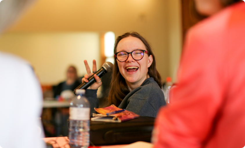 Audréanne Soenens from L'Arche Sudbury smiling at the Youth Leaders in-person course, holding up a peace sign and a microphone.