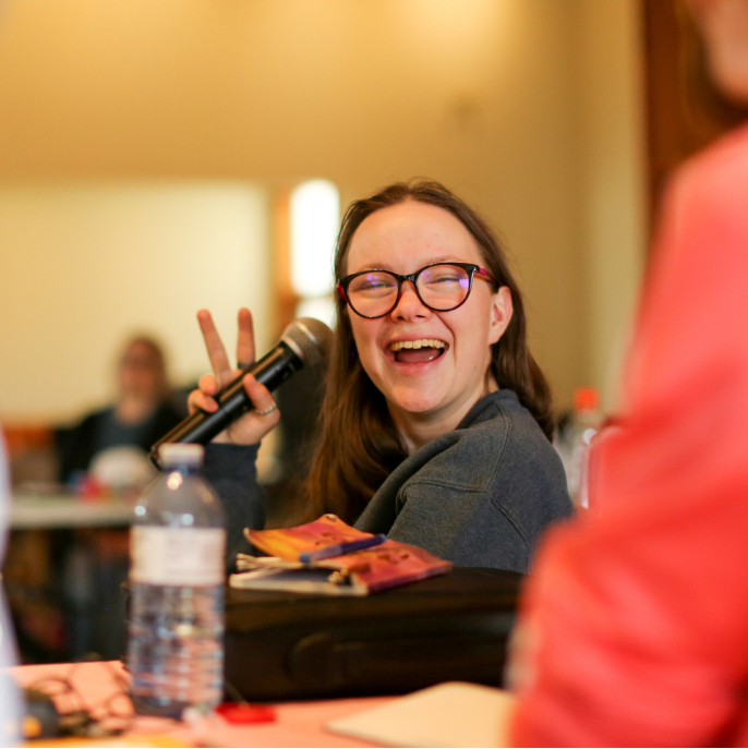 A young white woman with glasses laughing, holding up microphone and peace sign with her hands, at the Youth Leaders in person training.